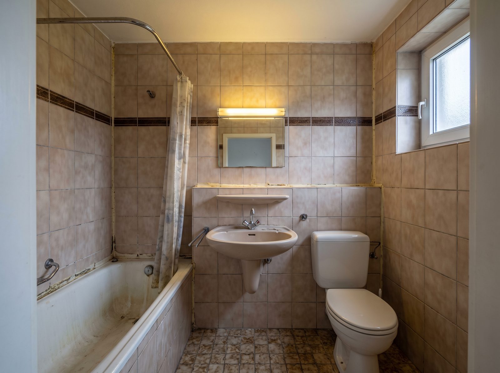 Dasselbe Badezimmer vor der Sanierung: beige-braune Fliesen mit Dekorbord&uuml;re, alte Wanne mit Duschvorhang, Standwaschbecken und vergilbte Fugen